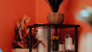 Ein kleines Holzregal mit einem gewebten Korb darauf, in dem eine Pflanze steht. Darunter befinden sich mehrere weiße Behälter und eine Vase mit Blumen.
