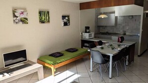 Ein kompakter Wohnraum mit Bett, Fernseher, Esstisch mit Stühlen, Küchenzeile und Bildern an den Wänden.