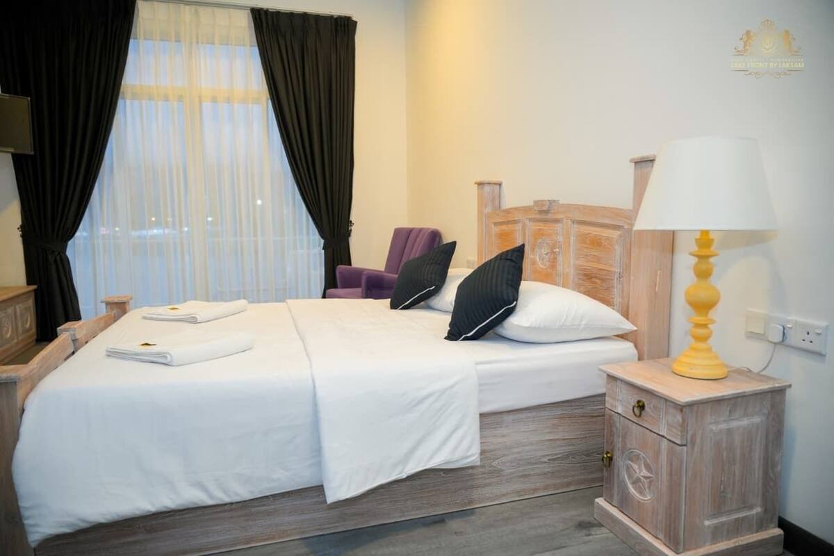 Un lit bien fait, avec du linge de lit blanc et des oreillers, une table de chevet en bois avec une lampe, et une chaise dans une pièce dotée d’une grande fenêtre et de rideaux.