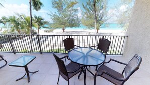 Een balkon met een glazen tafel en rieten stoelen, met uitzicht op een strand met palmbomen en een heldere lucht.