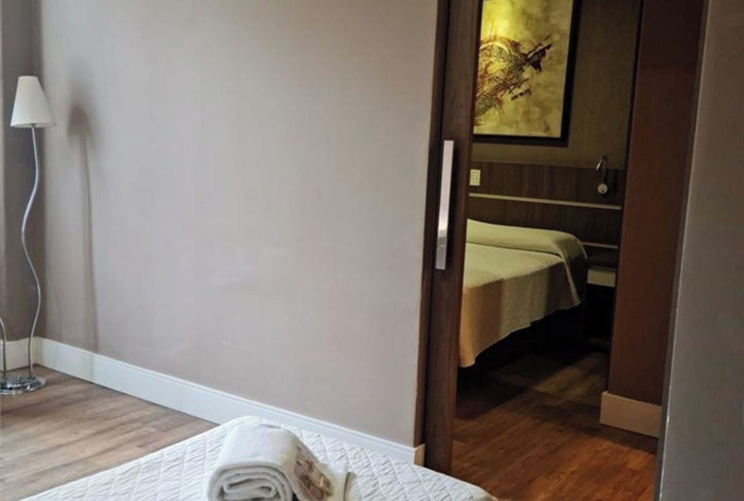 A hotel room with a wooden floor, a bed with a white comforter, a bedside lamp, and a painting on the wall.