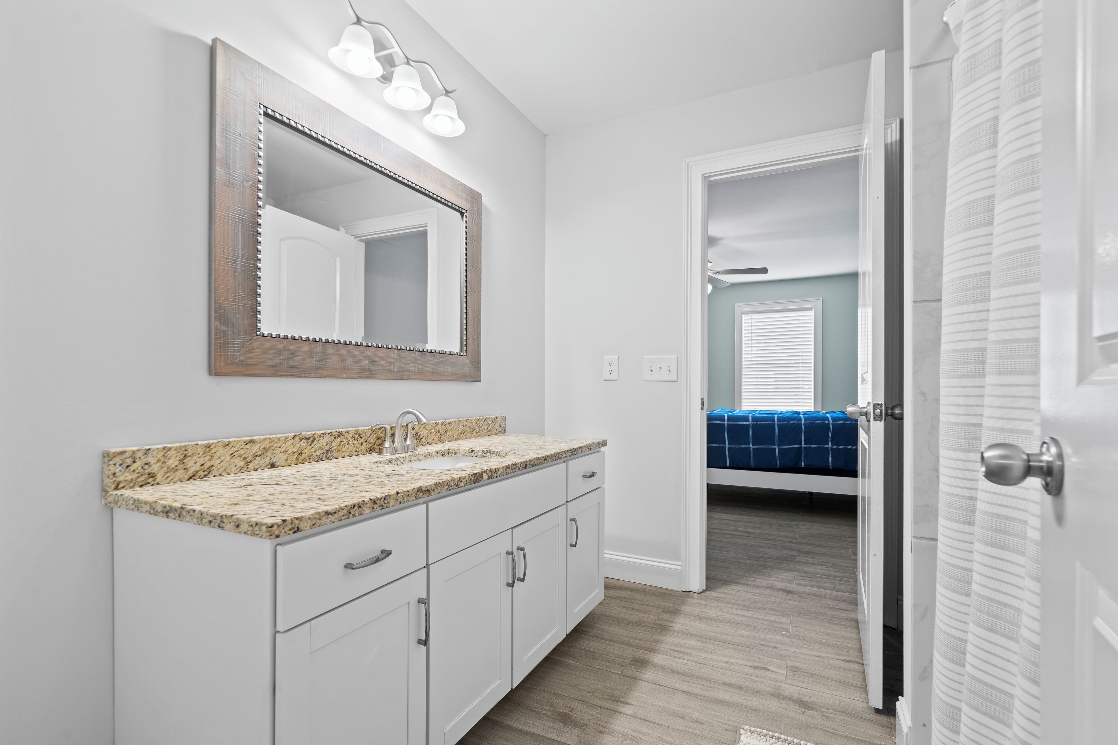 A bathroom with a granite countertop sink, a large mirror, and a door leading to a bedroom with a blue bedspread.