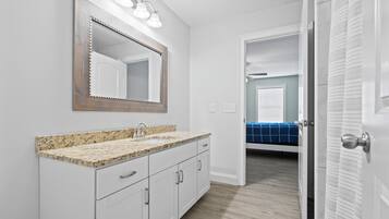 A bathroom with a granite countertop sink, a large mirror, and a door leading to a bedroom with a blue bedspread.