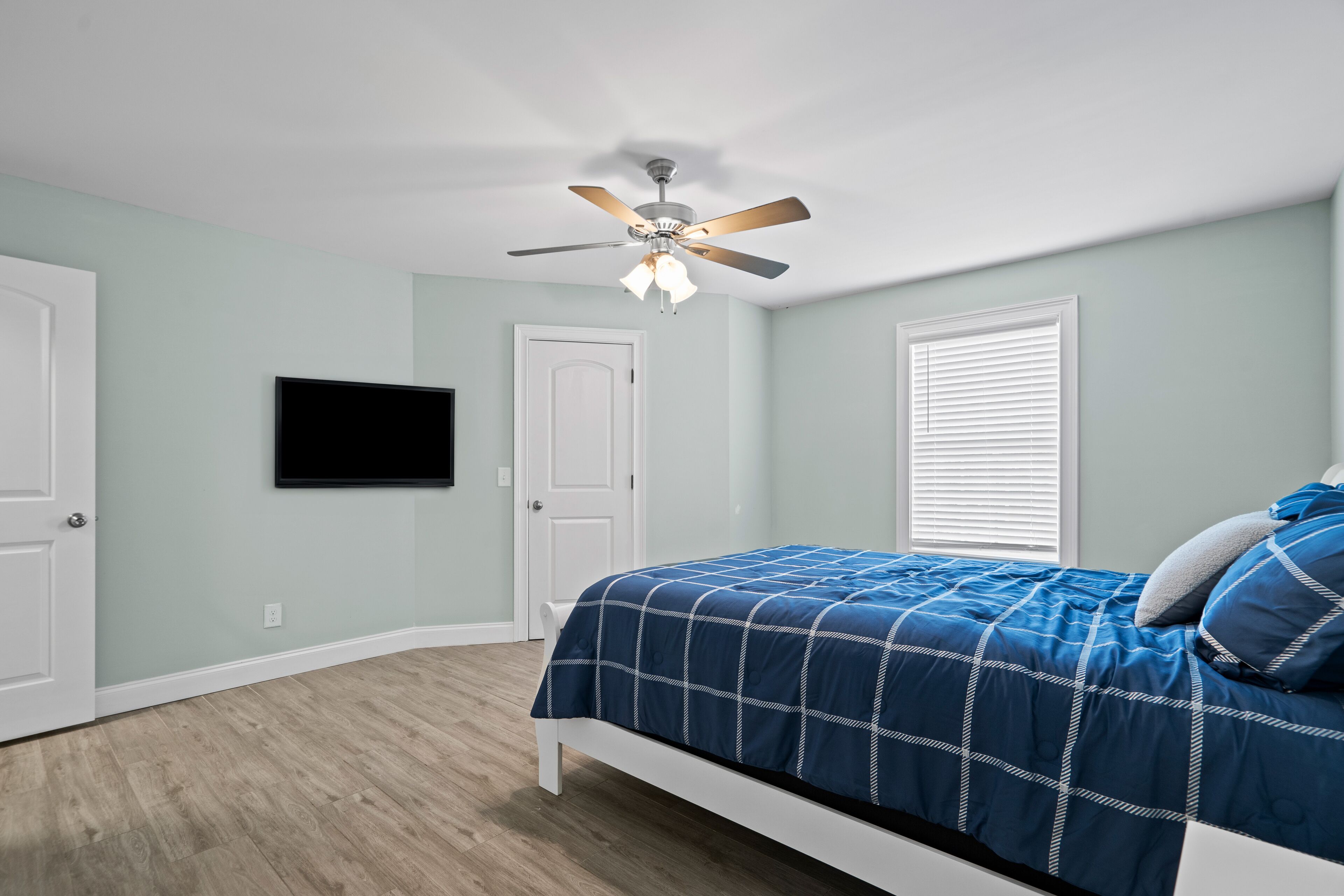 A bedroom with a bed, a ceiling fan, a TV mounted on the wall, and a window with blinds.