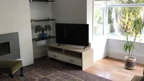 A living room with a flat-screen TV on a wooden stand, a bookshelf with books, a patterned rug, a green chair, and a window with a view of trees. - End terrace, sea views,short walk to the town and harbour of Ilfracombe (Ilfracombe)