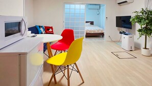 A modern, compact living space with a kitchenette, a dining area with colorful chairs, a bed, a TV, and a potted plant. - OLIVE stay (Fukuoka)