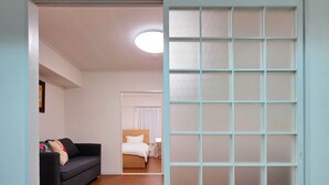 A room with a wooden floor, a dark-colored sofa, a bed with white bedding, and a frosted glass partition. - OLIVE stay (Fukuoka)