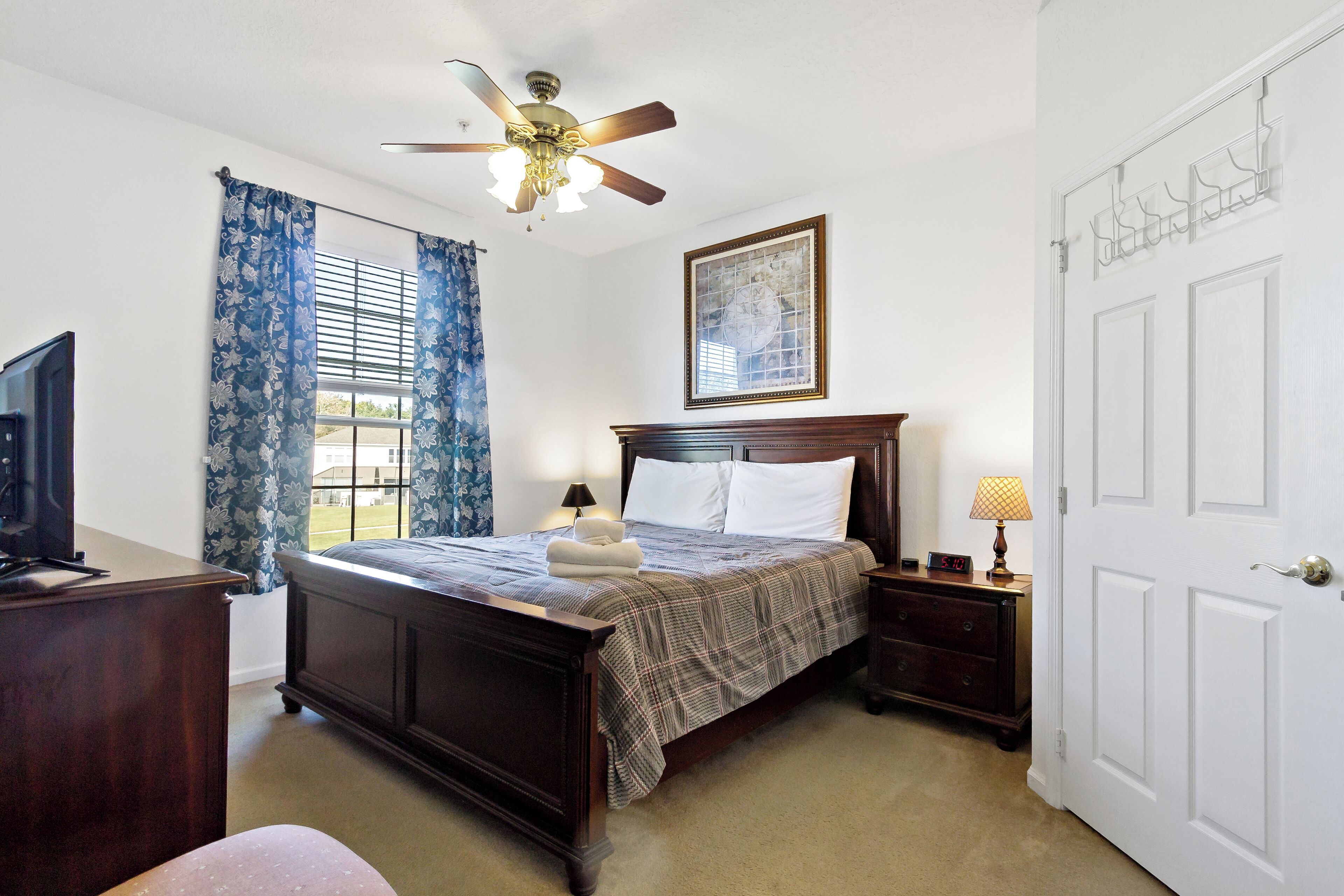 A bedroom with a wooden bed, a ceiling fan, a window with blue curtains, a bedside table with a lamp, and a door with a coat rack.