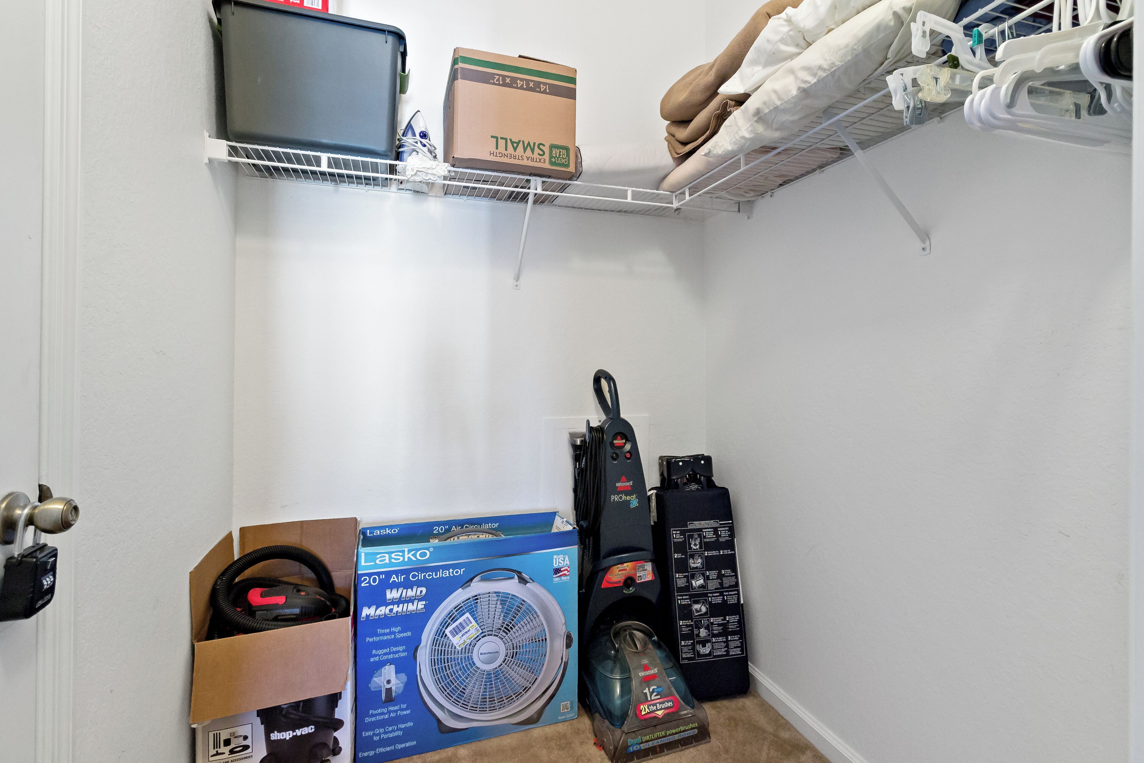 A storage area with a shelf holding a box, a vacuum cleaner, and a fan.
