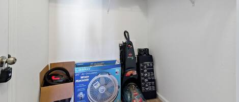 A storage area with a shelf holding a box, a vacuum cleaner, and a fan.