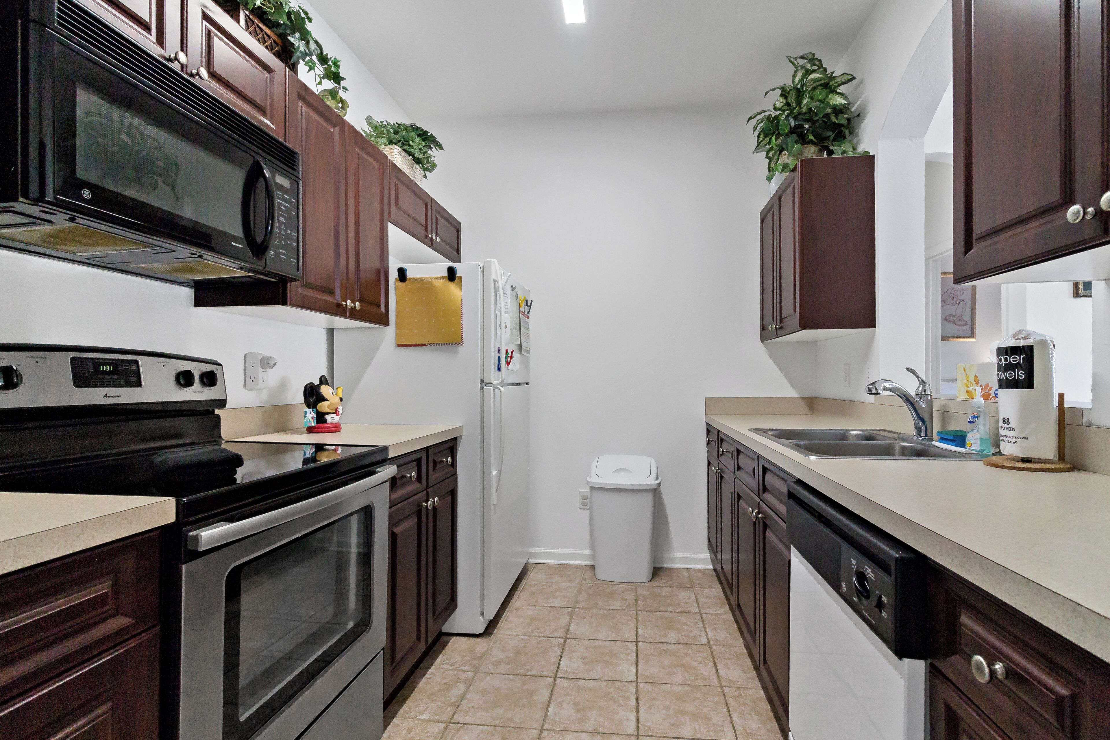 A kitchen with dark wooden cabinets, a stainless steel oven, a microwave, a refrigerator, and a dishwasher. There are two plants on the cabinets, and a Mickey Mouse figurine on the counter.