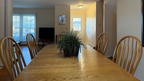 Ein Esszimmer mit einem langen Holztisch, sechs Holzstühlen, einem Blumentopf, einem Fernseher und einer Tür mit Fenster.