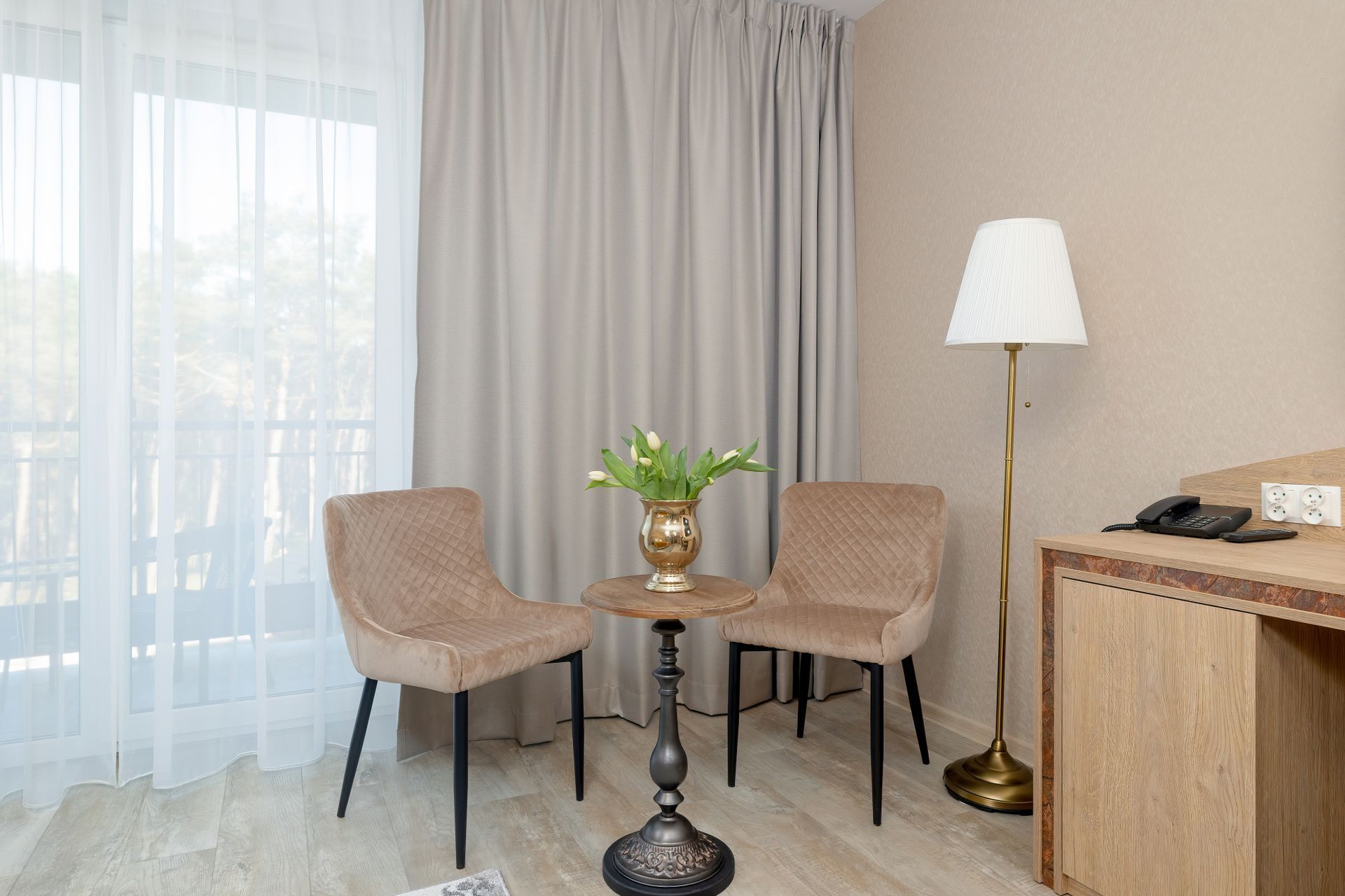 A hotel room with two chairs, a small table with a vase of flowers, a lamp, a desk with a phone, and a view through sheer curtains.