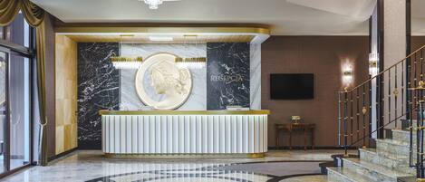 A reception area with a marble reception desk, a chandelier, and a staircase with a gold railing.