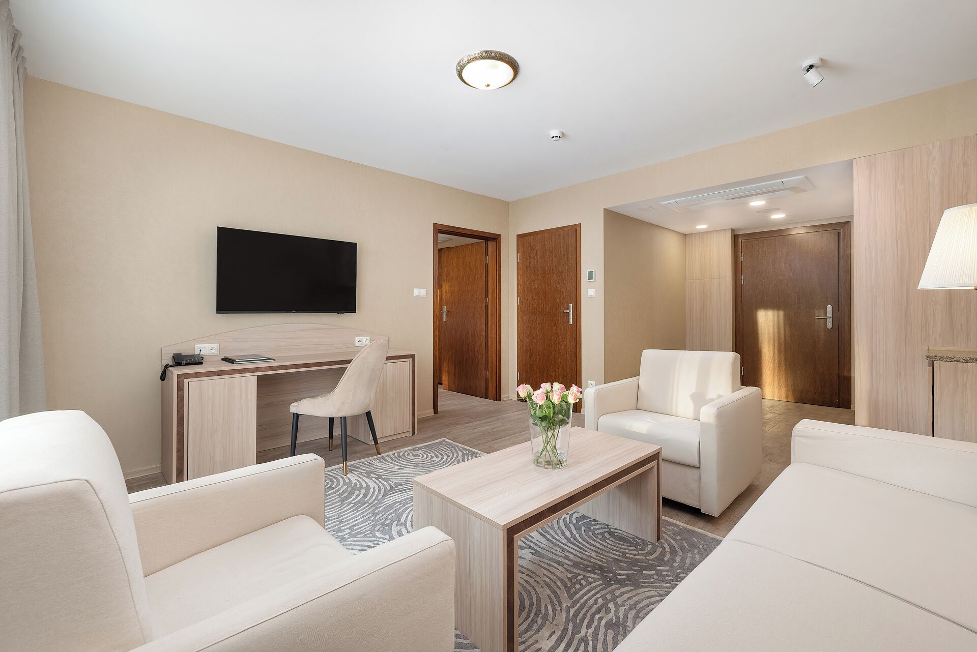 A modern hotel room with a flat-screen TV, a desk, two white sofas, a coffee table, and a vase with flowers.