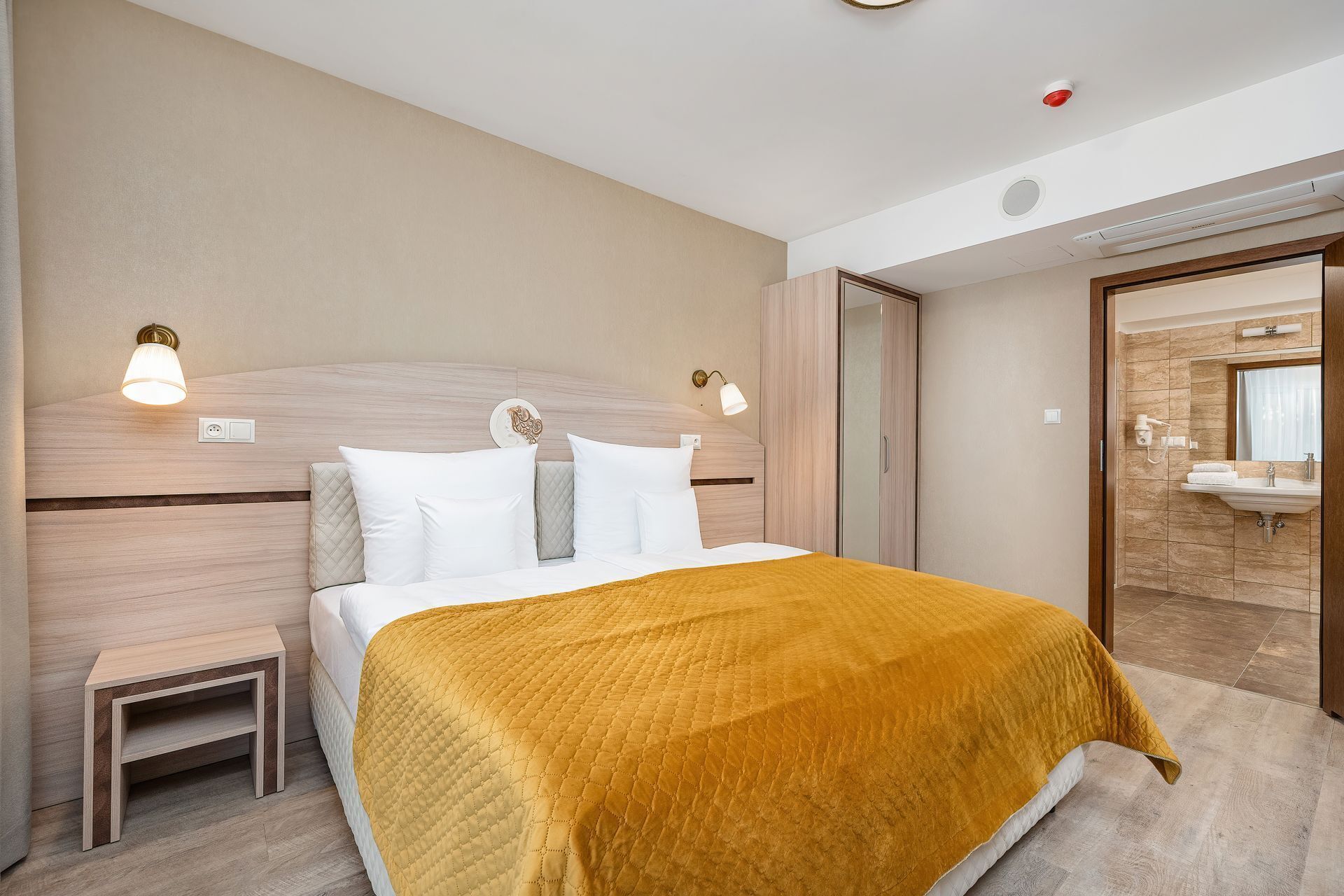 A hotel room with a bed, bedside tables, a bathroom with a sink and mirror, and a door leading to another room.