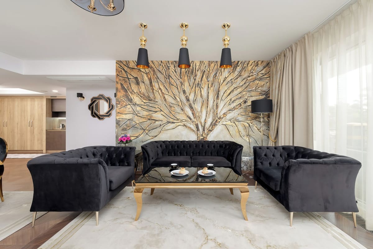Un salon moderne avec un ensemble de canapés noirs, une table basse en verre et une grande fresque murale représentant un arbre abstrait.