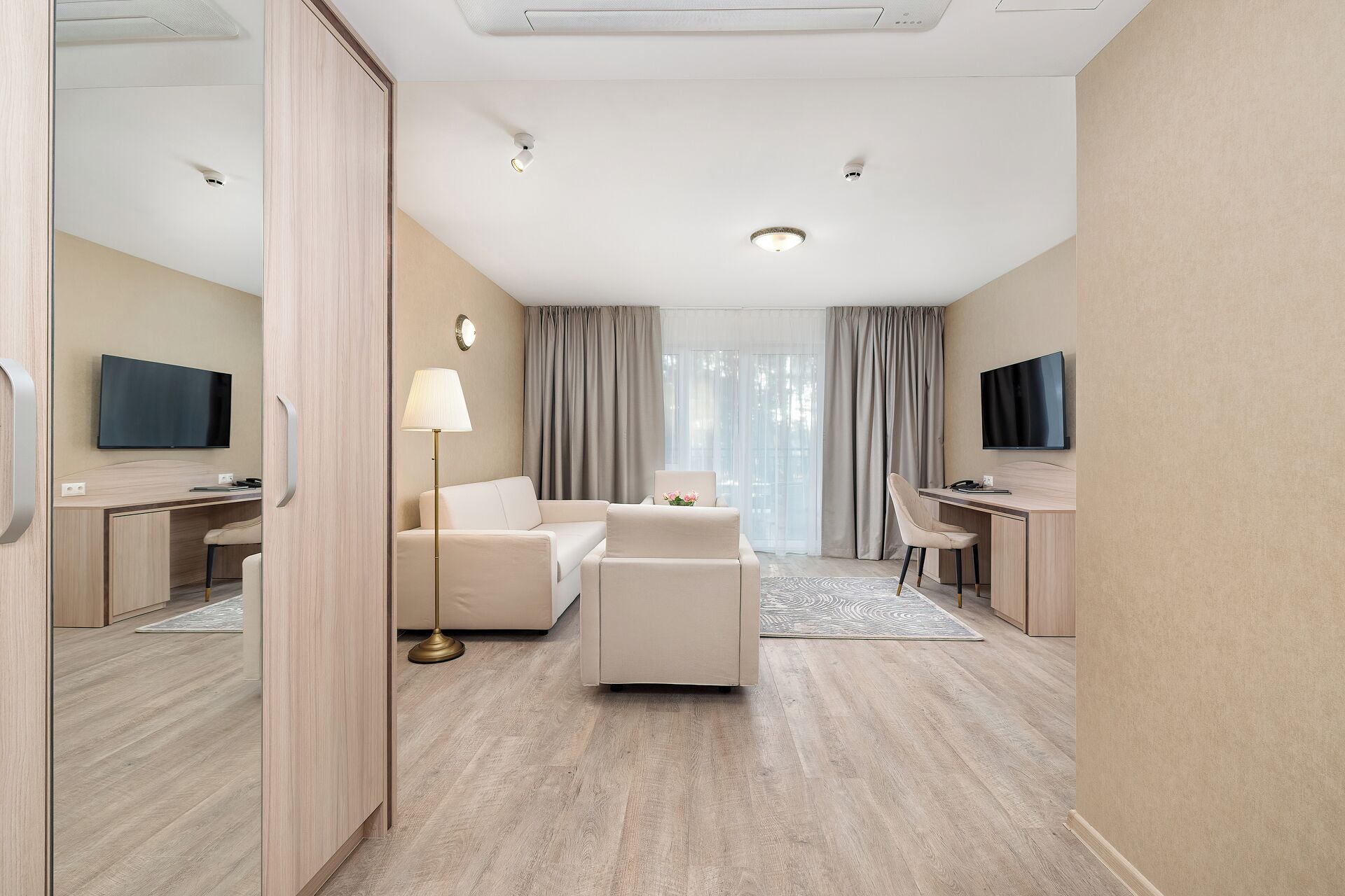 Una camera d'albergo moderna con un divano, due scrivanie, una TV e una lampada.