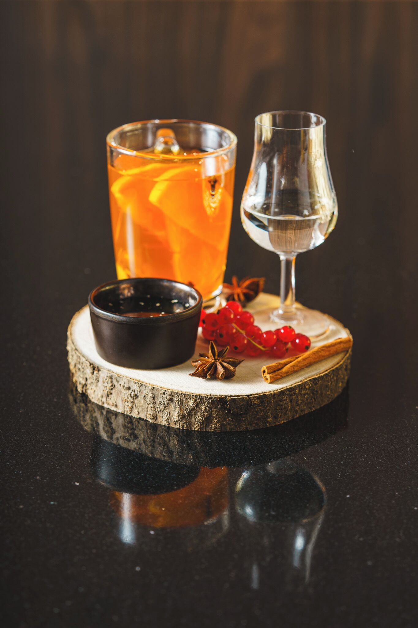 Ein Glas bernsteinfarbenes Getränk mit Eiswürfeln, eine kleine Schale dunkler Flüssigkeit und ein Glas klare Flüssigkeit auf einem Holzuntersetzer.
