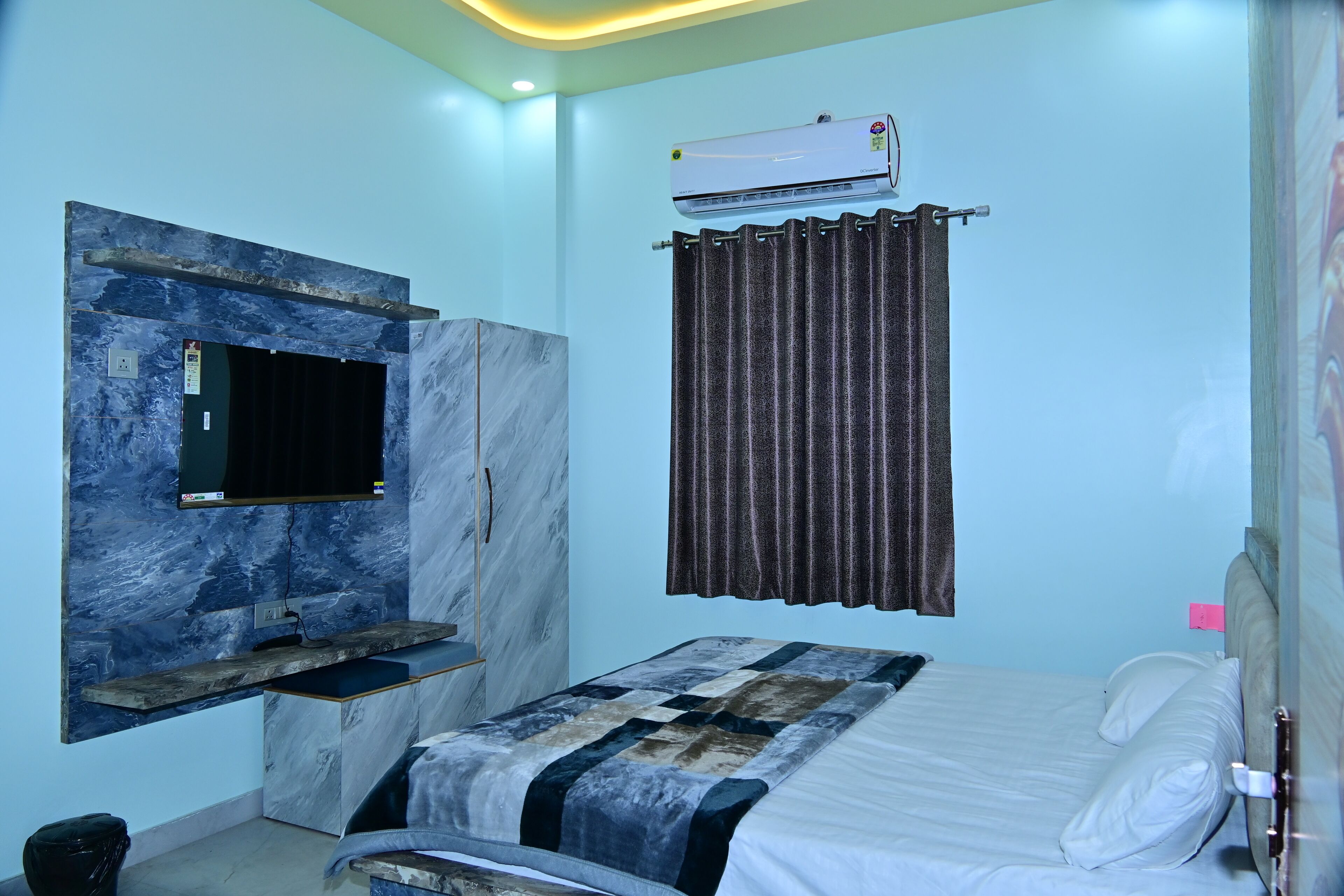 A bedroom with a bed, a TV mounted on a marble wall, an air conditioner, and a curtain.