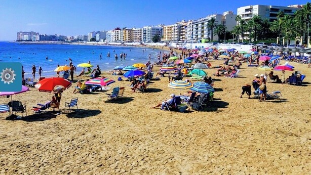 Ein Strand mit mehreren bunten Sonnenschirmen und Liegestühlen, einem klaren blauen Himmel und Gebäuden im Hintergrund.