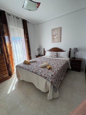 A bedroom with a large bed, two bedside tables with lamps, a wooden dresser, and a window with curtains. - Apartment on Avenue de la Mancha, across from the pool and a 15-minute walk to the beach (Torrevieja)