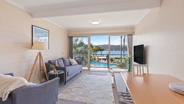 A modern living room with a sofa, a small table, a lamp, and a TV. There is a large window with a view of a pool and palm trees. - Fishpen Views - Merimbula (Merimbula)