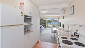 A kitchen with a stove, microwave, and cabinets. There is a dining area with a table and chairs, and a view of a pool and mountains in the background. - Fishpen Views - Merimbula (Merimbula)