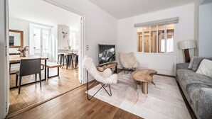 A modern living room with a wooden floor, a grey sofa, a white furry chair, and a wooden coffee table. There is a TV on a wooden stand and a painting on the wall. - Brina – Chamonix Apartment with Mont Blanc View (Chamonix-Mont-Blanc)