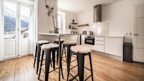 A modern kitchen with white cabinets, a stainless steel range hood, and a wooden floor. There are three bar stools with fluffy seats. A deer head decoration hangs on the wall, and a window offers a view of snow-capped mountains. - Brina – Chamonix Apartment with Mont Blanc View (Chamonix-Mont-Blanc)