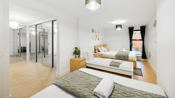 A modern hotel room with a large bed, a smaller bed, a desk, a chair, a plant, and a view of the city. - Huge Digbeth Apt, Sleeps 8, 5bed, Free Parking. (Birmingham)