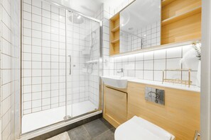 A modern bathroom with a glass shower enclosure, a white sink, and wooden cabinetry. - Huge Digbeth Apt, Sleeps 8, 5bed, Free Parking. (Birmingham)