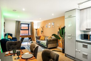 A modern living room with a sofa, dining table, chairs, and a kitchen area with cabinets and a microwave. - Huge Digbeth Apt, Sleeps 8, 5bed, Free Parking. (Birmingham)