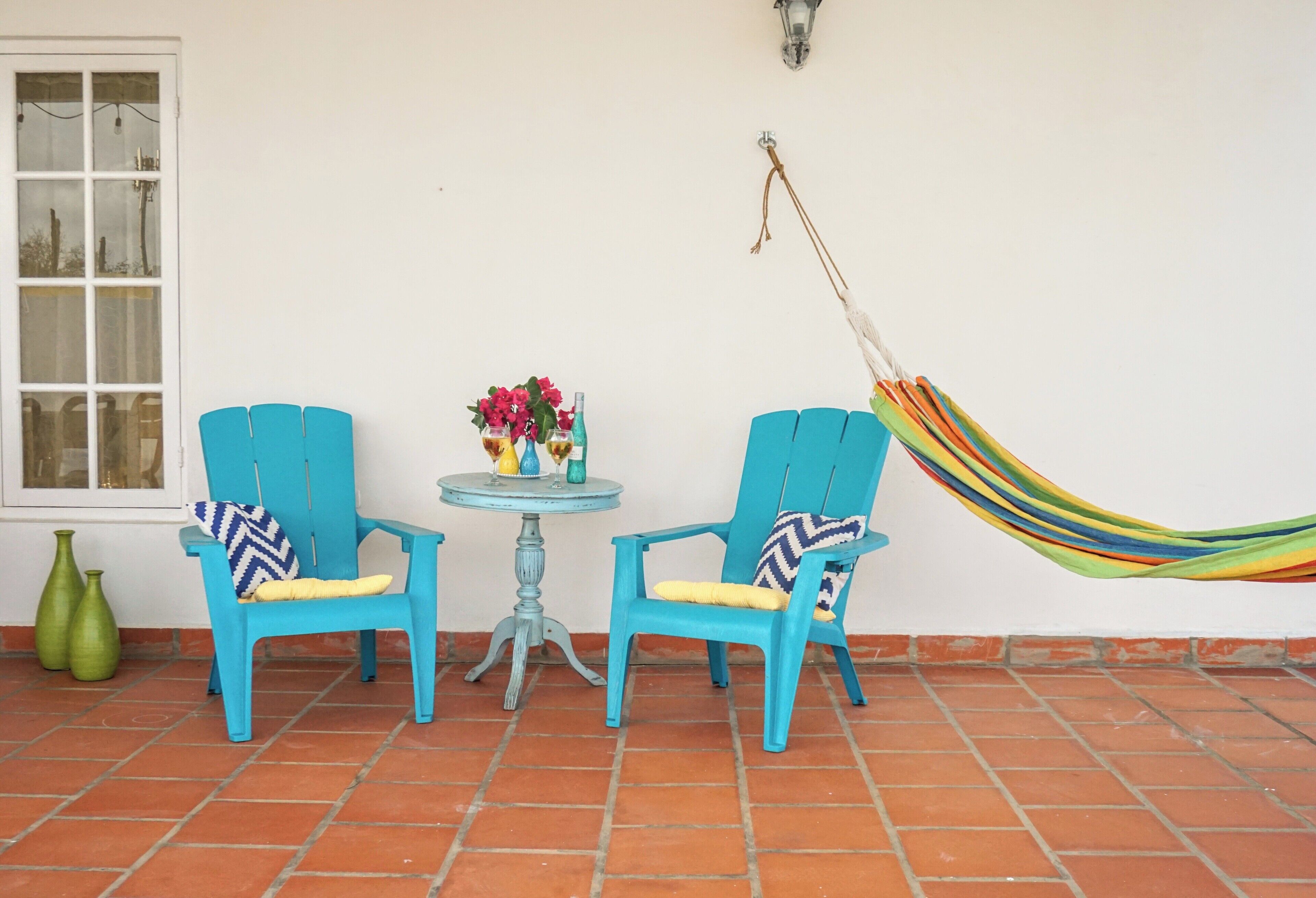 Two blue chairs with patterned cushions, a small round table with a vase of flowers, and a colorful hammock against a white wall.