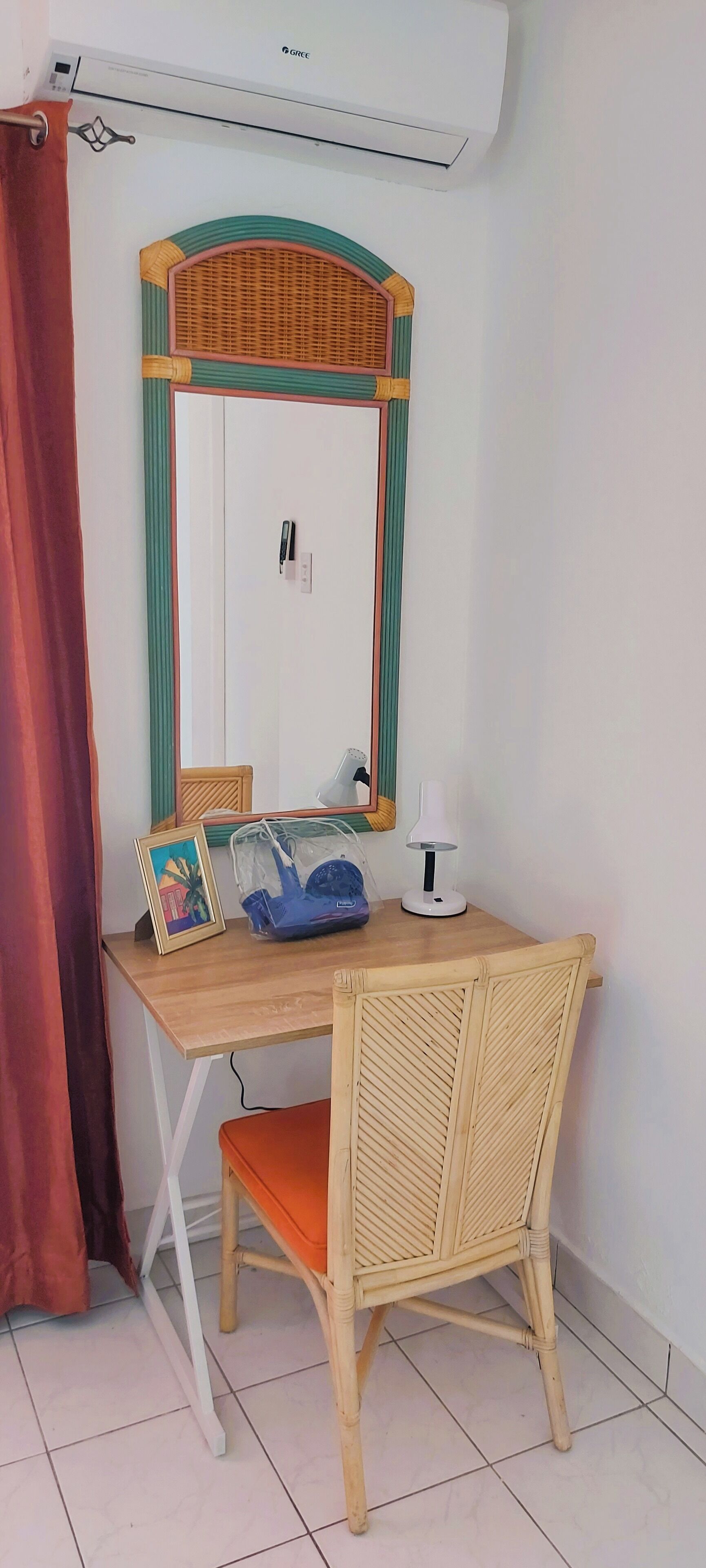 A room with a wooden desk, a wicker chair, a mirror with a green frame, a white air conditioner, and a red curtain.