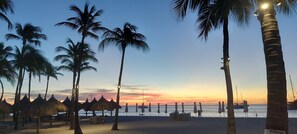 Une plage bordée de palmiers et ombragée par des parasols en chaume, sous un ciel au coucher du soleil.