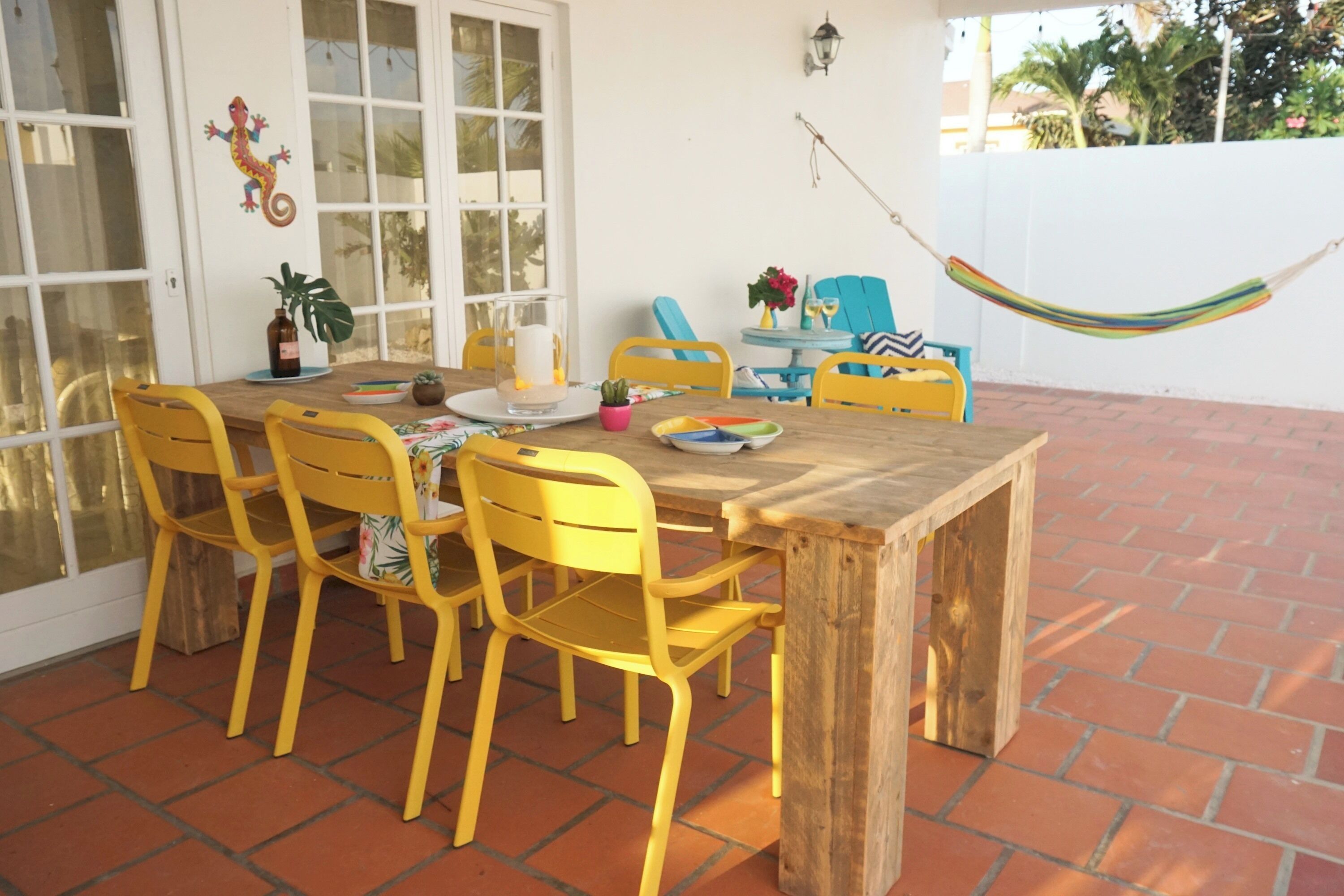 Eine Terrasse mit Holztisch und gelben Stühlen, einer Hängematte und einer weißen Wand mit einer dekorativen Eidechse.