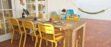 Eine Terrasse mit Holztisch und gelben Stühlen, einer Hängematte und einer weißen Wand mit einer dekorativen Eidechse.