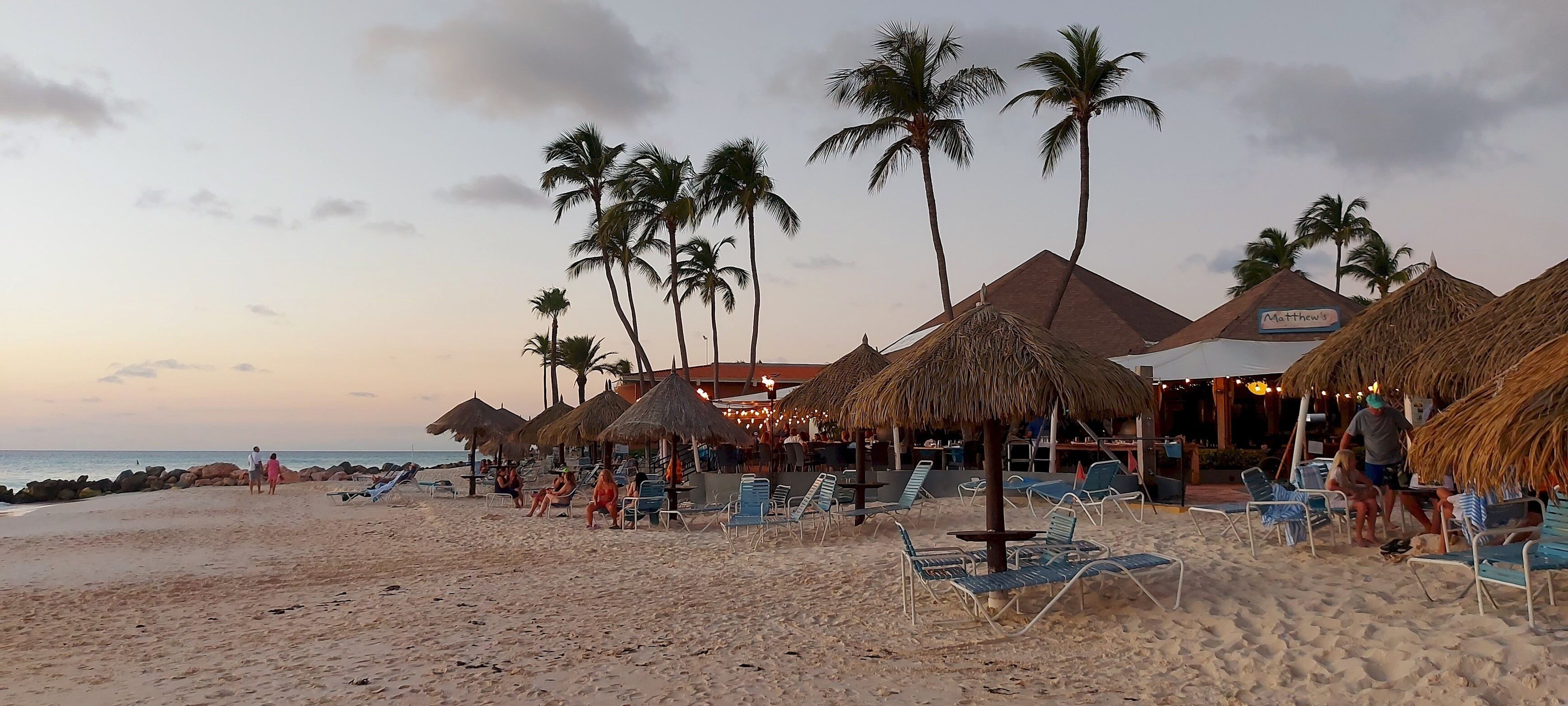 A beach with thatched-roof structures, palm trees, and lounge chairs.