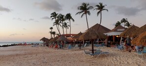 Une plage avec des constructions à toits de chaume, des palmiers et des chaises longues.