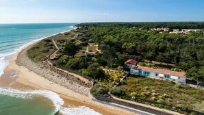 An aerial view of a coastal area with a beach, a house with a red roof, and surrounding greenery.