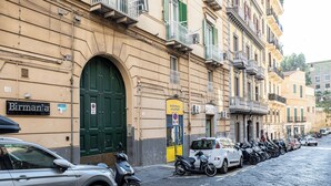 Une rue bordée de voitures et de motos garées, un immeuble avec une porte verte et une enseigne sur laquelle on peut lire « Birmana ».