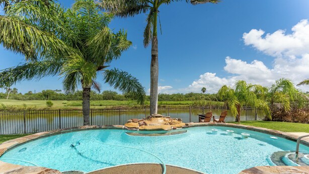 A swimming pool with a stone border, a small island with a hot tub, and a view of a lake and palm trees. - Poolside Bliss/fenced yard/water views/golf/pickleball/pets/tennis/sun & fun (Laguna Vista)