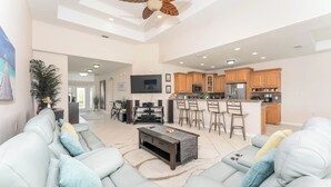 A spacious living area with a sofa, coffee table, and a ceiling fan. A kitchen with wooden cabinets and a bar area is visible in the background. - Poolside Bliss/fenced yard/water views/golf/pickleball/pets/tennis/sun & fun (Laguna Vista)