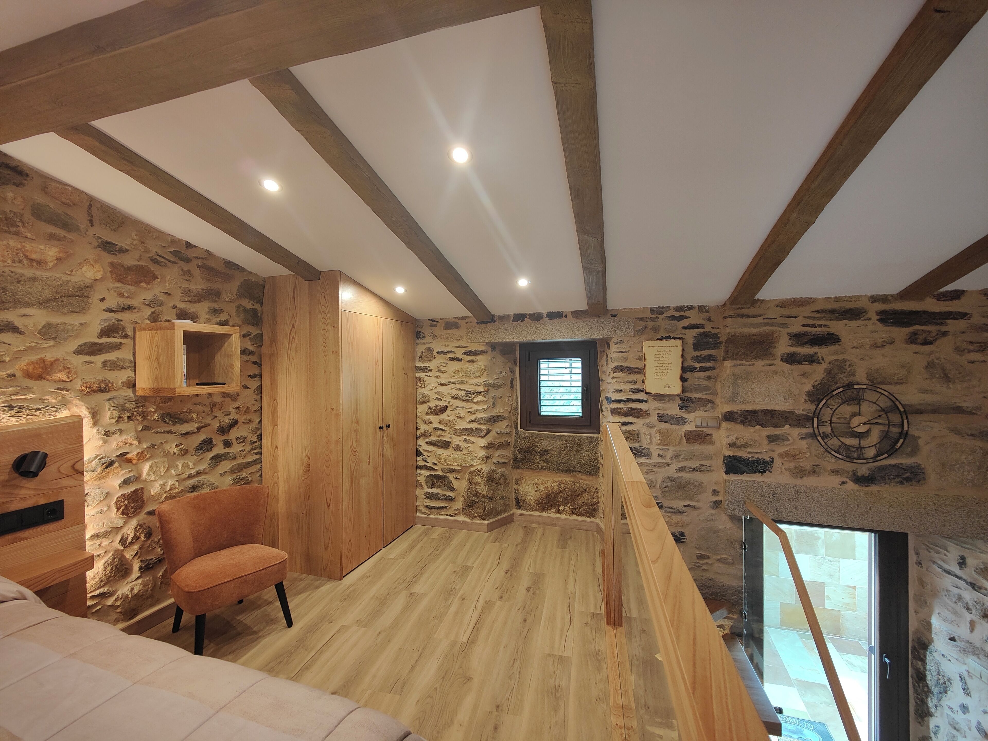 Een landelijke kamer met zichtbare stenen muren, houten balken, een houten garderobe, een comfortabele stoel en een klein raam.