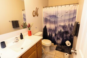 Une salle de bain avec un lavabo blanc, des toilettes, un rideau de douche orné d’un motif de forêt, un porte-serviettes et un miroir décoré d’un « M ».