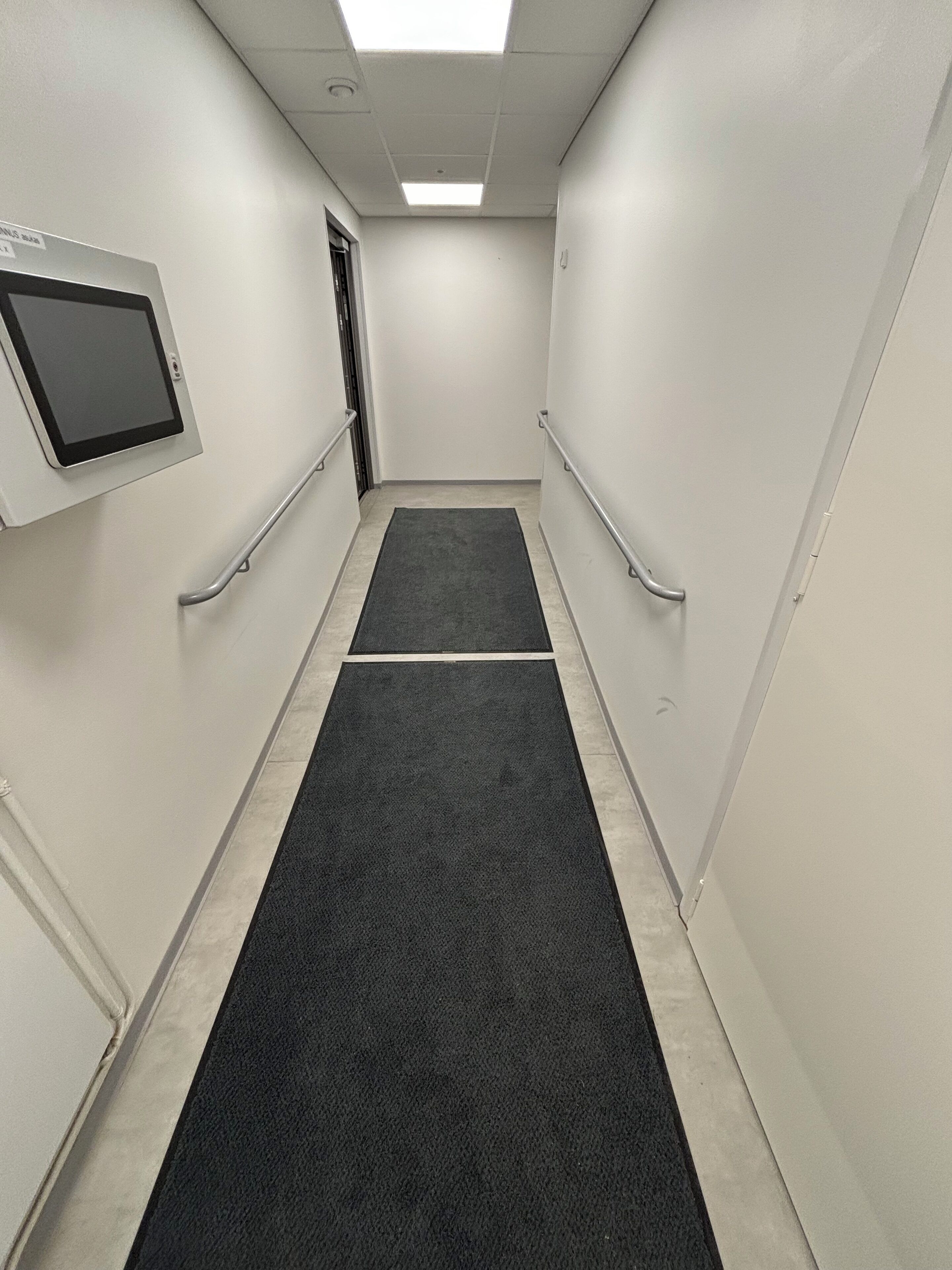 Un estrecho pasillo alfombrado, con paredes blancas, una lámpara en el techo y una puerta al final.