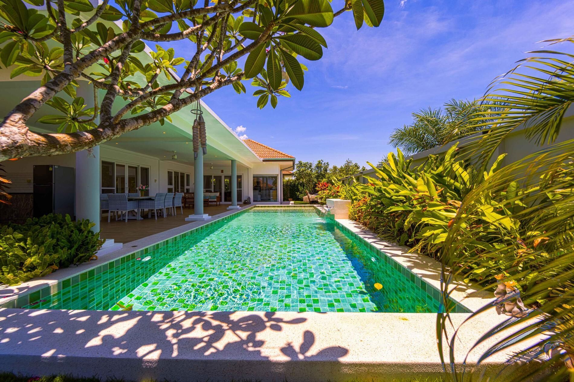 A swimming pool with mosaic tiles, surrounded by lush greenery and a covered patio area.
