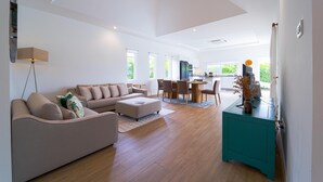 A spacious living area with a sofa, coffee table, and a dining table with chairs. There is a kitchen area with a refrigerator and a television. - TropicalFamilyVilla w Pool&Garden ML103 (Hua Hin)