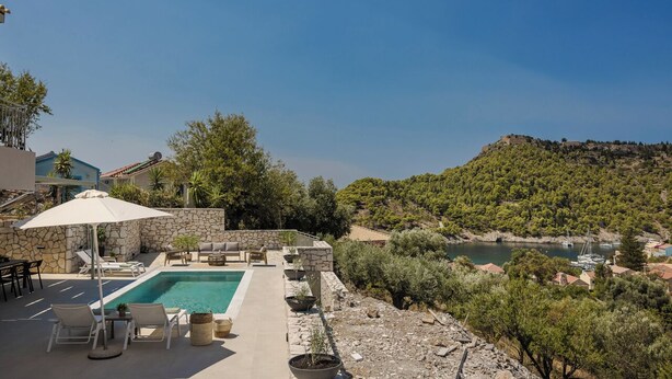Un espace piscine agrémenté de chaises longues et d’un mur en pierre, offrant une vue sur une baie où se trouvent des bateaux et un paysage vallonné.